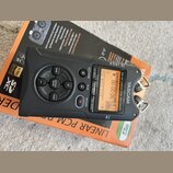 Černý digitální nahrávač TASCAM DR-40 LINEAR PCM RECORDER s displejem a ovládacími tlačítky. Zařízení leží na oranžovém originálním obalu. Na horní s…