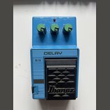 Modře barevný efekt pedál s nápisem DELAY a označením DL10. Na horní části jsou tři zelené oválné ovladače a nápisy OUT, DRY OUT, DC a IN. V dolní čá…
