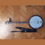 Na fotografii je banjo s bílou membránou a kovovým tělem, k němuž je přiložen černý popruh a malá krabička s příslušením. Na hmatníku jsou vidět orna…