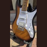 Elektrická kytara s tělem v provedení sunburst a bílým pickguardem. Nástroj má tři snímače typu single-coil, chromovaný most s páčkou a tři bílé ovlá…