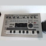 Stříbrný sekvenční syntezátor Roland MC-303 groovebox. Na předním panelu jsou umístěny ovládací knofíky, tlačítka a displej s nápisy jako CURRENT, NE…