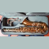 V otevřeném kufru značky Bam leží zlatý saxofon a dva zakřivené hrdly s korkem. Nástroj má lesklý povrch a je vybaven řadou klapek a mechanismů. Na s…