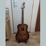 Akustická kytara s korpusem v tmavě hnědém přechodovém odstínu (sunburst) a hnedým hmatníkem. Na hlavě nástroje je viditelné logo Fender. Kytara stoj…