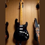 Černá elektrická kytara s hmatníkem ze světlého dřeva. Na hlavě je viditelné logo Fender. Kytara má tři snímače (dvě humbuckery a jeden jednosnímač),…