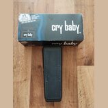 Na fotografii je černý wah pedál s gumovou texturovanou povrchovou vrstvou, umístěný před svou originální krabicí. Na krabici je bílý nápis cry baby.…
