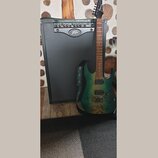 Na fotografii je elektrická kytara opřená o kytarový kombo zesilovač. Kytara má zelený gradientní lak s viditým zrnitím dřeva a na hlavě je bílý nápi…