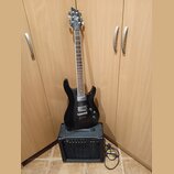 Černá elektrická kytara s dvojstupňovými humbuckery, opřená o černý kytarový kombo zesilovač. Na hlavě kytary je bílý nápis Cort. Zesilovač má na pře…