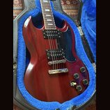 Kytara s červeným lakem a černým plechtrátem, umístěná v otevřeném kufru s modrou výplní. Nástroj má dva černé humbuckery, chromovaný most a čtyři če…