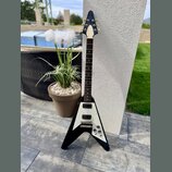 Černá elektrická kytara s V-tvarým korpusem a bílým pickguardem. Na hlavě hmatníku je vidět nápis Aria. Nástroj má dva humbuckery, tři ovládací knofl…