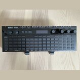 Černý polysequencer s nápisy KORG a SQ-64 POLY SEQUENCER. Zařízení obsahuje mřížku šedých tlačítek, několik otočných ovladačů a malý displej. Na horn…