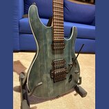 Elektrická kytara s modrozeleným průsvitným povrchem, pod kterým je vidět výrazná struktura dřeva. Nástroj má bílý lem kolem korpusu, dva humbuckery…