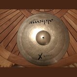 Kovový činel v přirozené zlatavé barvě s viditelným rýhováním. V horní části je černý nápis „alchemy cymbals“. Ve spodní části je černý kříž a vedle…