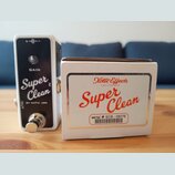 Bílá efekt pedál s černým předním panelem a kovovým spínacím tlačítkem. Na panelu jsou nápisy GAIN, Super Clean a BY xotic usa. Vedle pedálu je bílá…