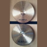 Dvě mosazné činely s viditelným broušením povrchu. Na obou je uveden nápis „istanbul Agop Handmade cymbals from Turkey“. Spodní činel má v dolní část…