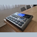 Šedý elektronický bicí modul s modrým displejem a velkým ovládacím knoflíkem. Na horní části je viditelné logo Roland. Zařízení obsahuje množství gum…