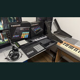 Sada hudebních zařízení a příslušenství na stole. Jsou zde dvě krabice s nápisy MASCHINE PRODUCTION AND PERFORMANCE SYSTEM a MASCHINE JAM PRODUCTION…