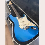 Modrá elektrická kytara typu Stratocaster s krémovým pickguardem a hnedým hmatníkem, umístěná v otevřeném tvrdém kufru. Na hlavě krku je viditelné lo…