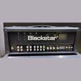 Černý kytarový zesilovač s kovovou mřížkou, na které je bílý nápis Blackstar. Přední panel obsahuje řadu ovládacích knoflíků a přepínačů s popisky ja…