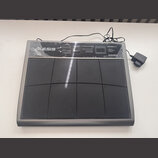 Šedý MIDI kontrolér s osmi černými gumovými pady. V levém horním rohu je bílé logo ALESIS a v pravém horním rohu nápisy ControlPad, USB/MIDI Percussi…