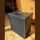Černý kombozesilovač s texturovaným povrchem a integrovanou rukojetí. Na vnitřní straně rukojeti je žlutý nápis "The Acoustic People". Na boční stěně…