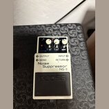 Bílý efekt pedál s černým spínacím tlačítkem. Na zařízení jsou nápisy BOSS, Noise Suppressor NS-2 a popisky ovládacích prvků REDUCTION, MODE, OUTPUT,…