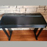 Černý dřevěný stolek pro klavír nebo digitální piano s výraznou strukturou žilování. Má obdélníkový tvar s mírně vyčnívající přední deskou a dvěma ši…