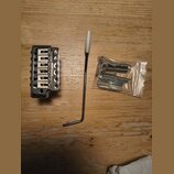 Kytarový kobylník stříbrné barvy s nápisem Floyd Rose. Vedle něj je umístěna páka s bílým koncem a plastový sáček s montážním příslušenstvím, včetně…