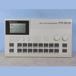 Bílý rytmický automat s displejem a ovládacími tlačítky. Na horní části je nápis RHYTHM COMPOSER TR-505 a v levém horním rohu logo Roland. Zařízení o…