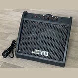 Černý kytarový kombo zesilovač s kovovou mřížkou včelíkového vzoru. Na přední straně je umístěno bílé logo JOYO. Horní panel obsahuje červený vypínač…