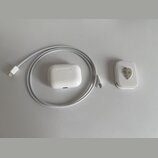 Bílé nabíjecí pouzdro k bezdrátovým sluchatkům, šedý pletený kabel s konektory USB-C a bílý síťový adaptér. Na přední straně pouzdra je vidět malá ko…