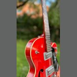 Červená polodublová elektrická kytara s bílým lemováním hran. Na těle jsou vidět dva stříbrné humbuckery a černý plecháč. Povrch laku je lesklý, bez…