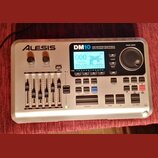 Stříbrný elektronický bicí modul značky ALESIS, model DM10. Na přední straně je displej s nápisem Fusion a Tempo 24 BPM, pět posuvníků pro ovládání h…