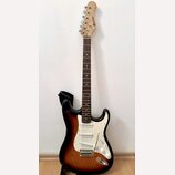 Elektrická kytara s tłem v provedení sunburst a bílým pickguardem. Na hlavě hředu je viditelné logo "Squier". Nástroj má tři snímače typu single-coil…