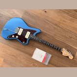 Modrá elektrická kytara s tmavě hnědým vzorovaným pickguardem a hnědým hmatníkem. Na hlavě nástroje je logo Fender a nápis JAZZMASTER®. Kytara leží n…