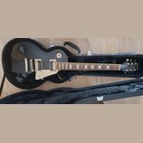 Černá elektrická kytara s tvarem single-cut v otevřeném černém kufru. Na hlavě je bílý nápis Epiphone a stylizovaný nápis Les Paul. Kytara má dva hum…