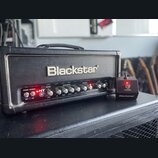 Černý kytarový zesilovač s nápisem Blackstar na předním panelu. Pod logem se nachází ovládací prvky s nápisy POWER, STANDBY, HT 5, REVERB (LEVEL), EQ…
