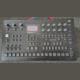 Černý analogový tabletop syntetizér s nápisem Analog Four MKII. Přístroj obsahuje displej, množství tlačítek, posuvníky a otočné knoflíky. V pravém h…