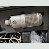 Stříbrný kondenzátorový mikrofon v černé pěnové výplni kufru. Na těle je vidět logo značky a nápis NEUMANN MICROPHONE. V dolní části kufru jsou vidět…
