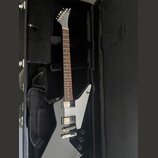 Šedá elektrická kytara s hranatým tělem a bílým pickguardem, položená v černém kufru. Nástroj má dva humbuckery a tři ovládací knoflíky. Na hlavě kyt…