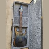 Elektrická kytara s černým gradientním povrchem a viditelnou strukturou dřeva na horní části těla. Na hlavě je bílý nápis Ibanez. Nástroj je umístěn…