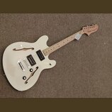 Bílá polodublová elektrická kytara s přírodním hřidlem a hlavou. Na hlavě je nápis "Squier STARSCASTER". Tělo má dva humbuckery, dva ovládací knoflík…