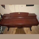 Hnědé dřevěné piano s uzavřenou klaviaturní víkem. V horní části je umístěno zlaté logo s nečitelným nápisem. Nástroj stojí na dvou černých nohách a…