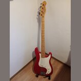 Červená elektrická basová kytara s bílým pickguardem a hřednou z jasného dřeva. Na hlavě je nápis "Fender Jazz Bass". Nástroj vykazuje běžné známky p…