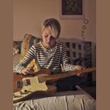Dítě v proužkovaném tričku drží v rukou elektrickou kytaru s přírodním dřevěným povrchem těla. Kytara má bílý pickguard, dva snímače a tři ovládací k…