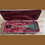Černá elektrická kytara v hliníkovém kufru s červeným plyšovým vnitřkem. Na hlavě nástroje je bílý nápis Ibanez. Kytara má dva humbuckery, tremolo mo…