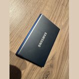 Na fotografii je modrý obdélníkový předmět s kovovým povrchem, který leží na dřevěném stole. Uprostřed je bílý nápis SAMSUNG. Povrch předmětu vypadá…
