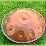 Měděný handpan s několika vyhlubenými tóny, položený na zelené trávě. Na povrchu je bílý nápis „Harmony Handpan“ a pod ním logo s textem „Seld the ha…