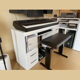 Černý digitální klavír umístěný na bílém stole s přihrádkami. Na levé straně klavíru je nápis MONTREUX. K sestavě patří černá polstrovaná piano stoli…