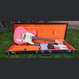 Růžová elektrická kytara s bílým pickguardem v otevřeném černém kufru s oranžovou výstelkou. Na hlavě kytary je viditelné logo Fender. V kufru jsou v…
