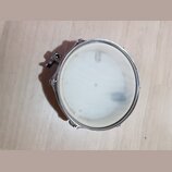 Kruhlový bicí buben s bílým průsvitným blánou a chromovaným obručem. Na levé straně je vidět kovový mechanismus pro uchycení. Povrch blány vykazuje m…